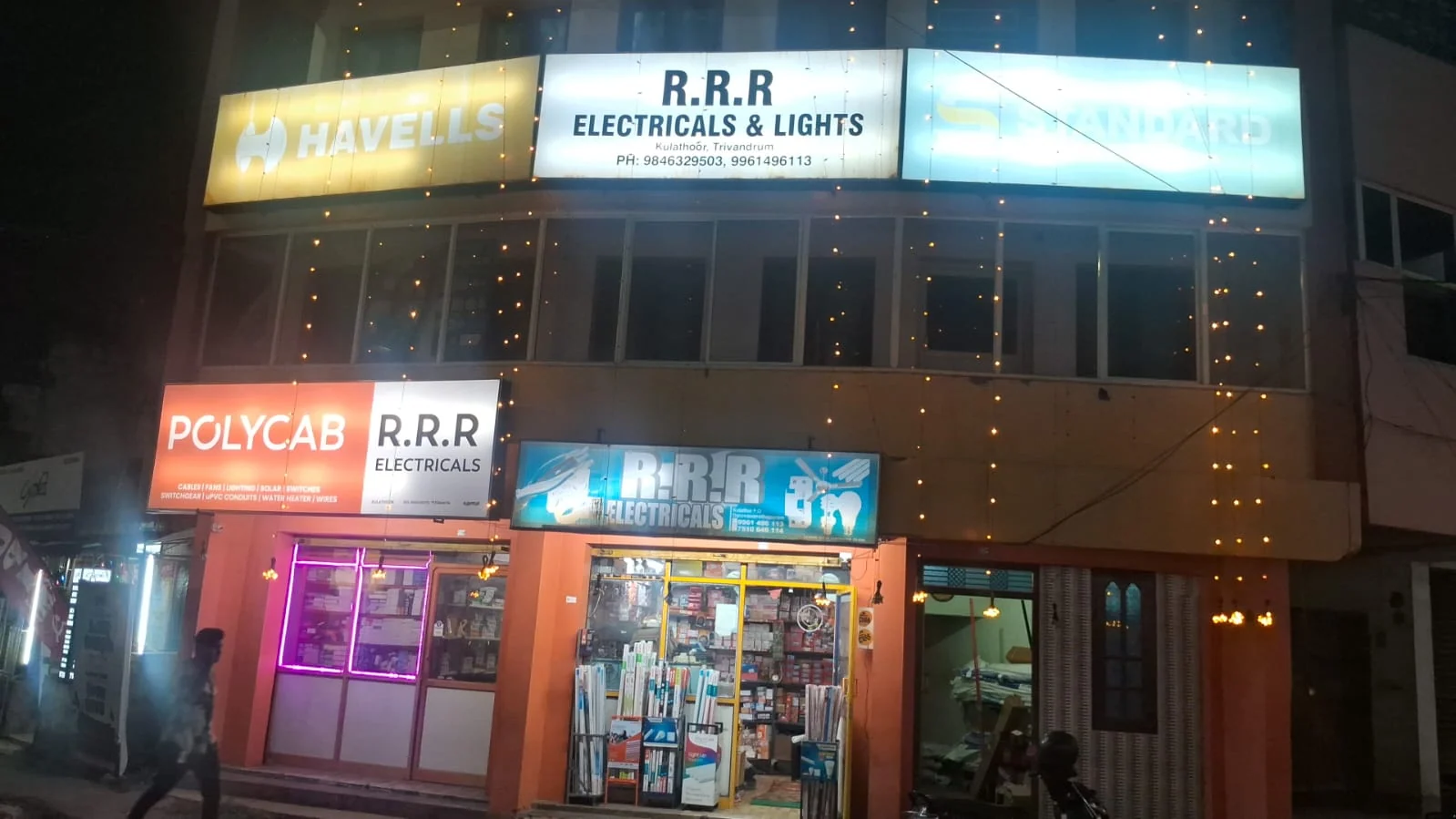 The actual RRR Electricals and Lights building at night, with its illuminated signs and existing string lights